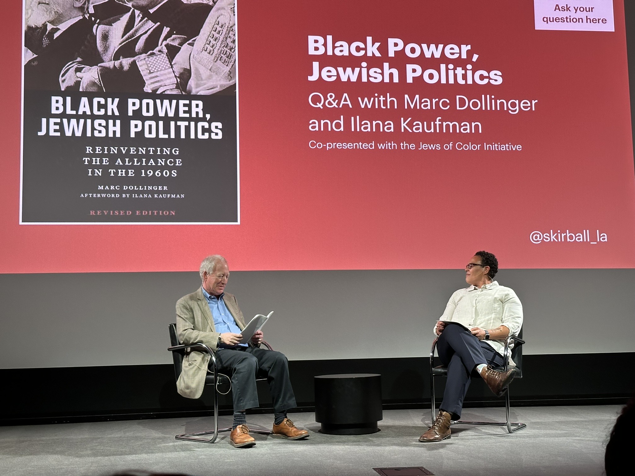 Marc Dollinger and Ilana Kaufman sit comfortably on chairs on a stage in front of a very large screen displaying a presentation slide with the following text: Black Power, Jewish Politics, Q&A with Marc Dollinger and Ilana Kaufman, co-presented with the Jews of Color Initiative. The background is a strong bright red and the slide includes an image of the cover of the book